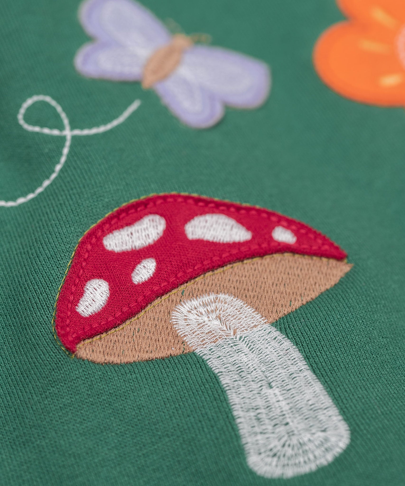 A close up of the Frugi kids pippa holly green fun guys pinafore outfit showing the mushroom applique detail on green organic cotton fabric.