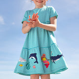 A smiling young child wearing the Frugi Pixy Sea Moss Mini Stripe Dress, with a Sealife print along the bottom, featuring whales, dolphins, sharks, a Mermaid and some small fish, surrounded by colorful seaweed, stood outside.