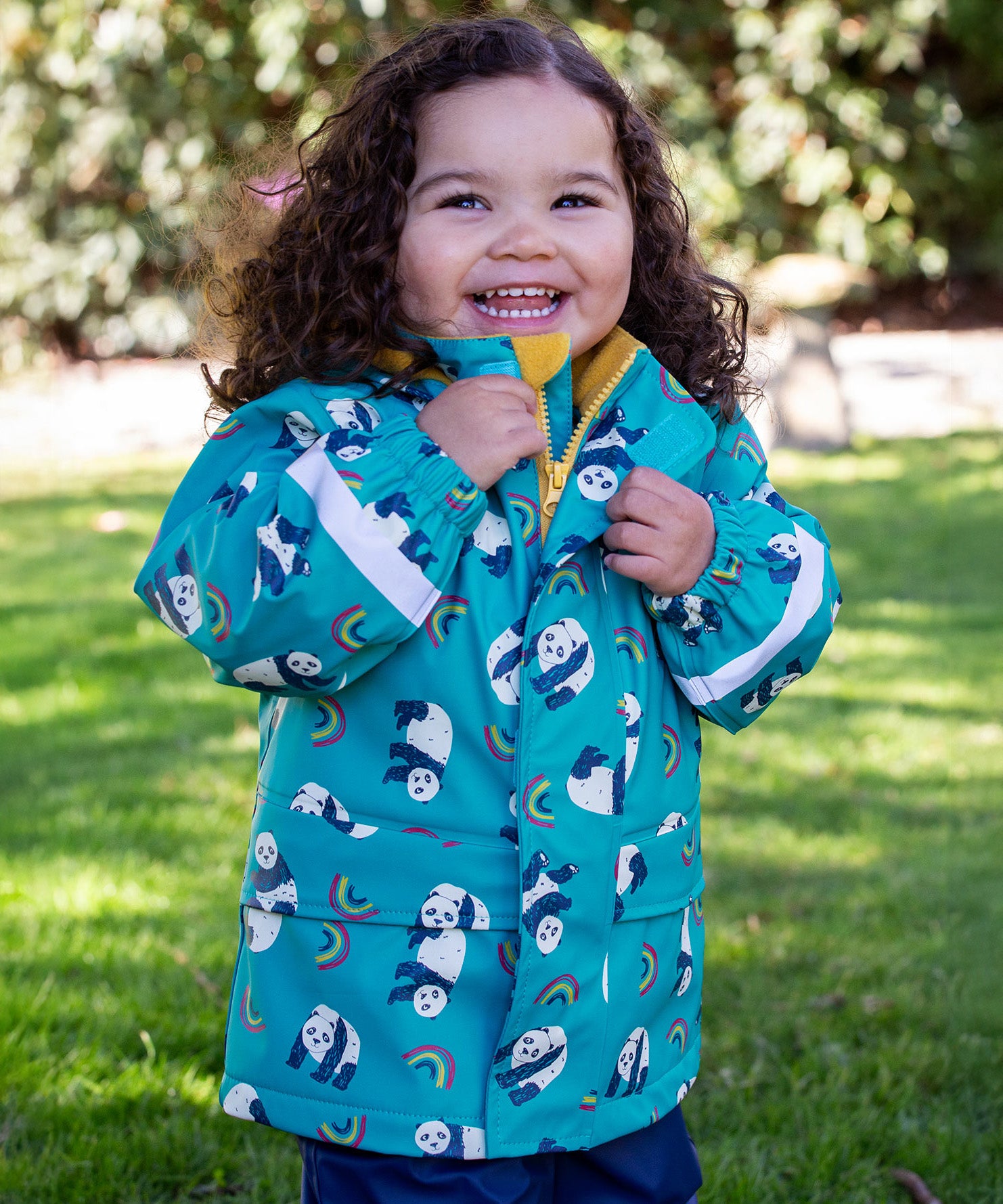 A toddler standing outside and wearing the Frugi puddle buster panda play coat.   light blue waterproof hooded coat with playful prints of pandas and rainbows with yellow zip trim