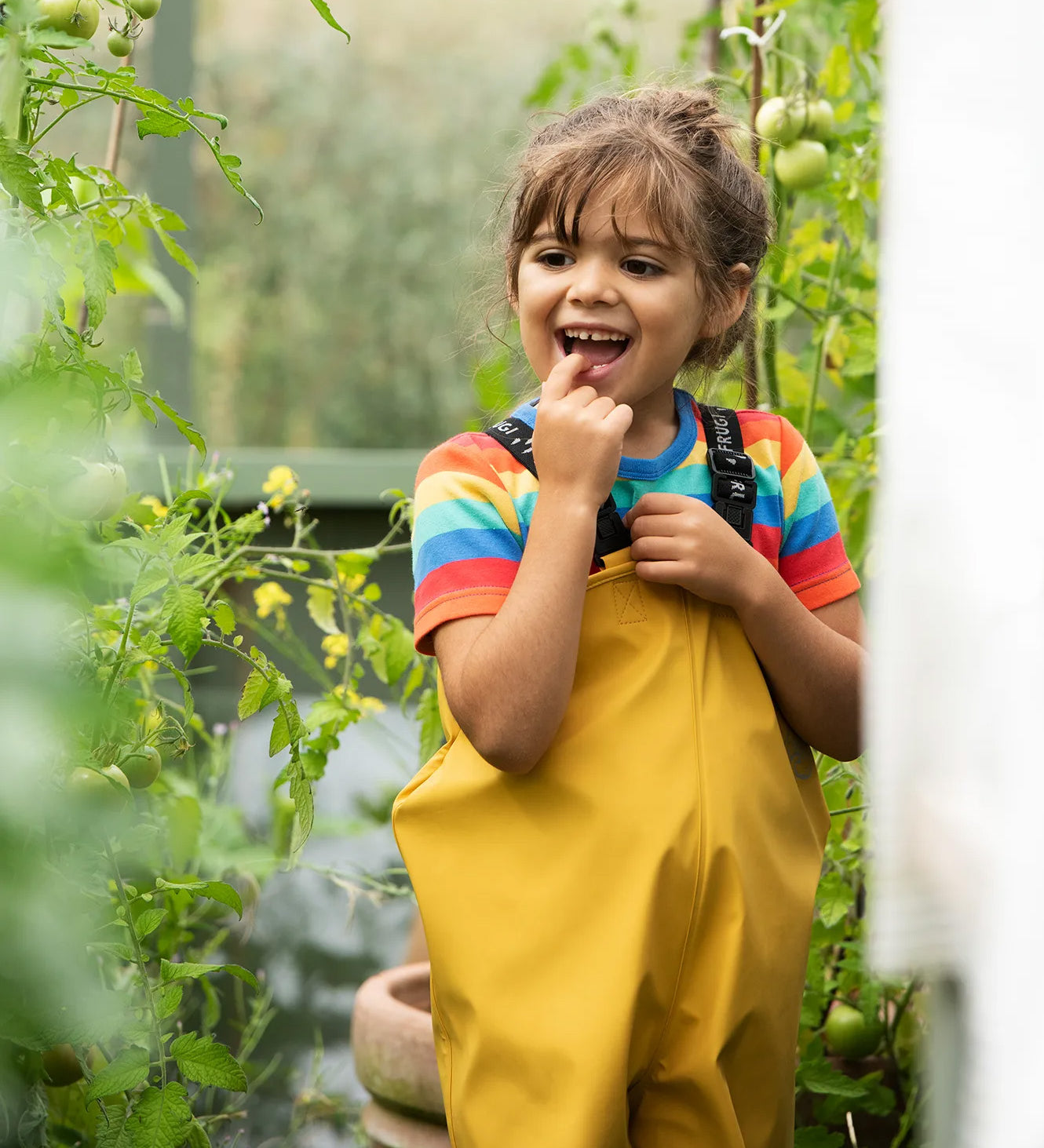 A little girl wearing Frugi waterproof Puddle Buster trousers in a garden, they're made from recycled plastic bottles and come in bright yellow