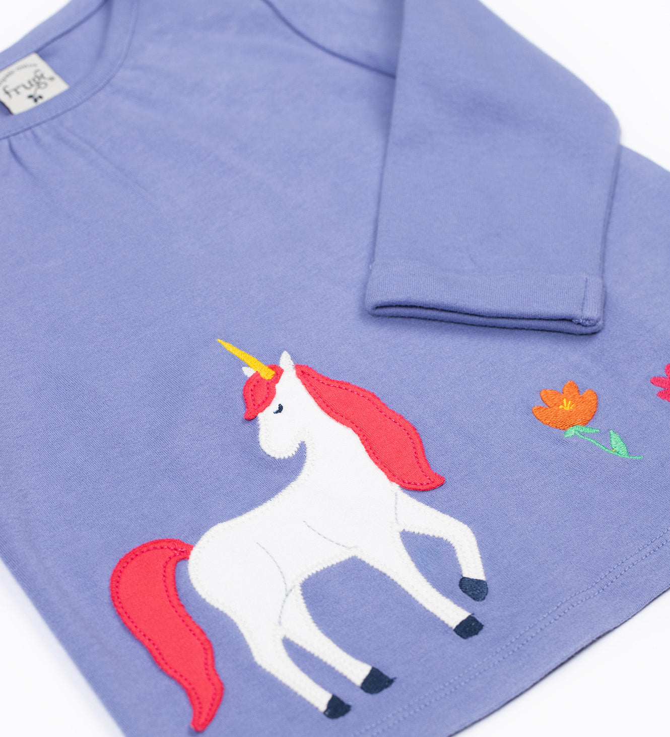 A close up of the unicorn applique  on the front of the Frugi grape purple unicorn Organic Cotton Ottlie Outfit. An outfit with a purple coloured top with a unicorn applique on the front and pink unicorn print leggings. This outfit is a part of a wide range of organic cotton clothing for children available here at Babipur.
