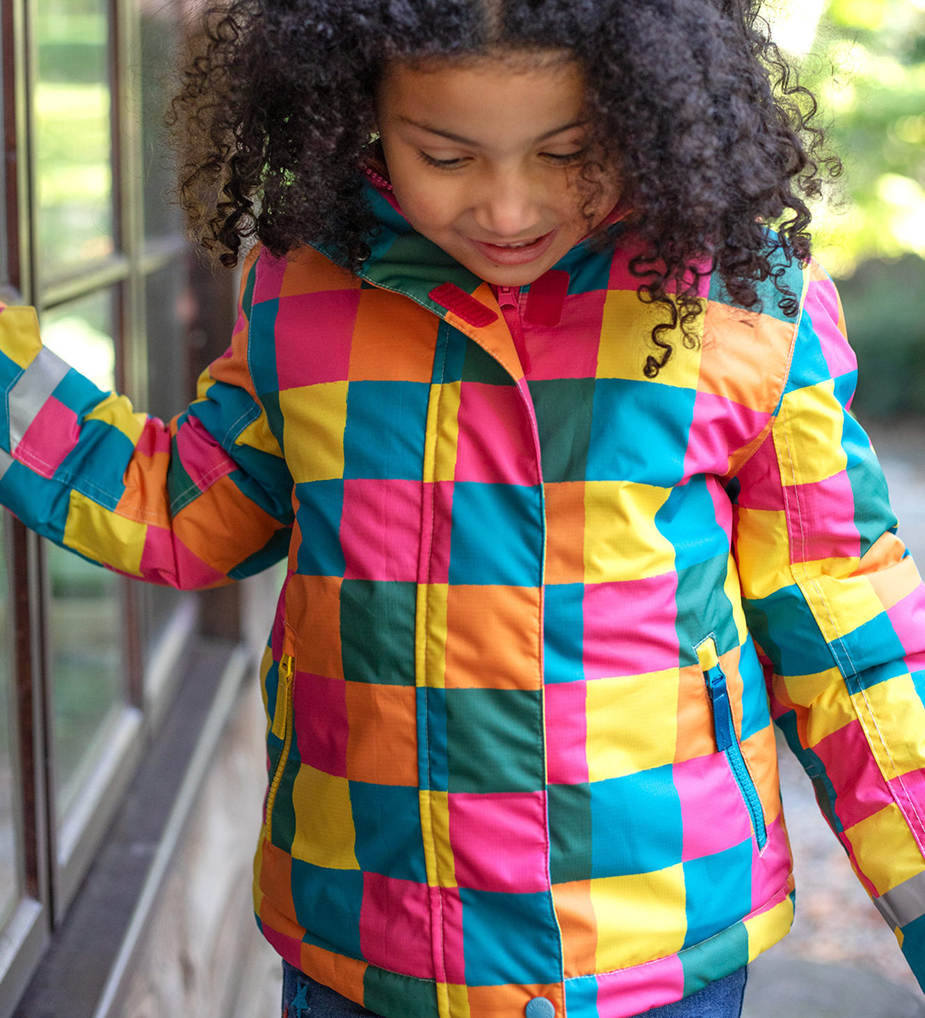 A child wearing the Frugi kids snow and ski coat in rainbow check. A multicoloured check winter coat with 2 side pockets with reflective strips on the arms
