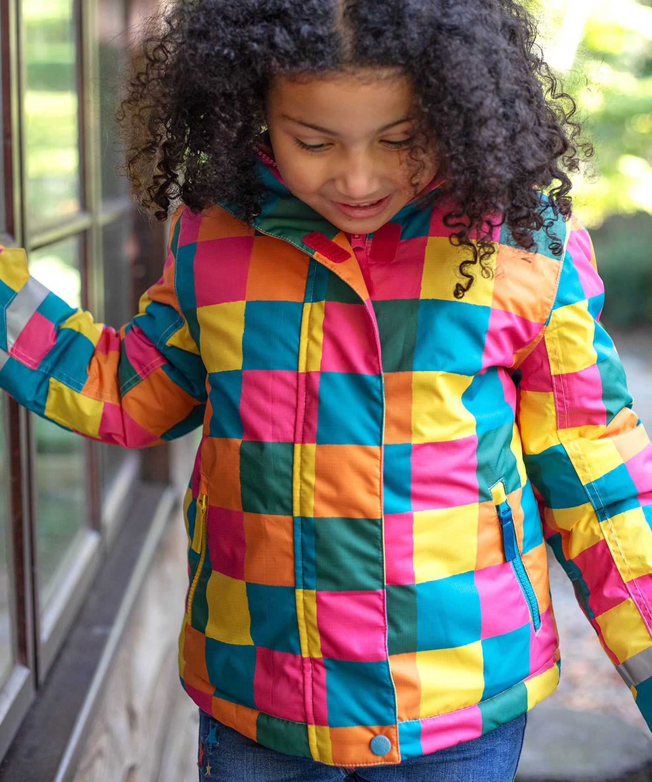 A child wearing the Frugi kids snow and ski coat in rainbow check. A multicoloured check winter coat with 2 side pockets with reflective strips on the arms