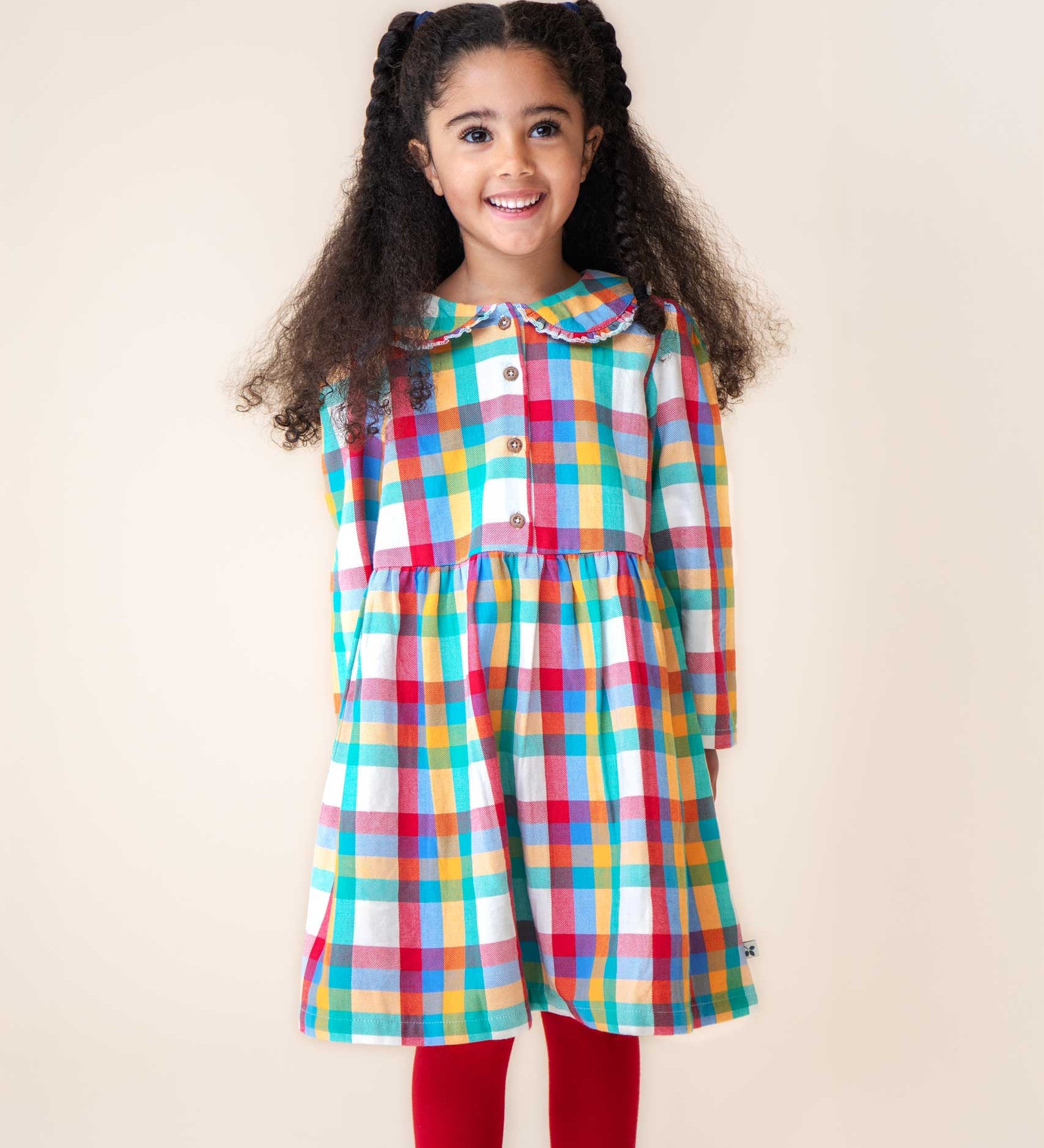 A child wearing the Frugi Rainbow Check Organic Cotton Ffion Dress. The dress has a rounded collar details and button closure on the front. This festive kids dress is a part of a wide range of organic cotton clothing for children available here at Babipur.