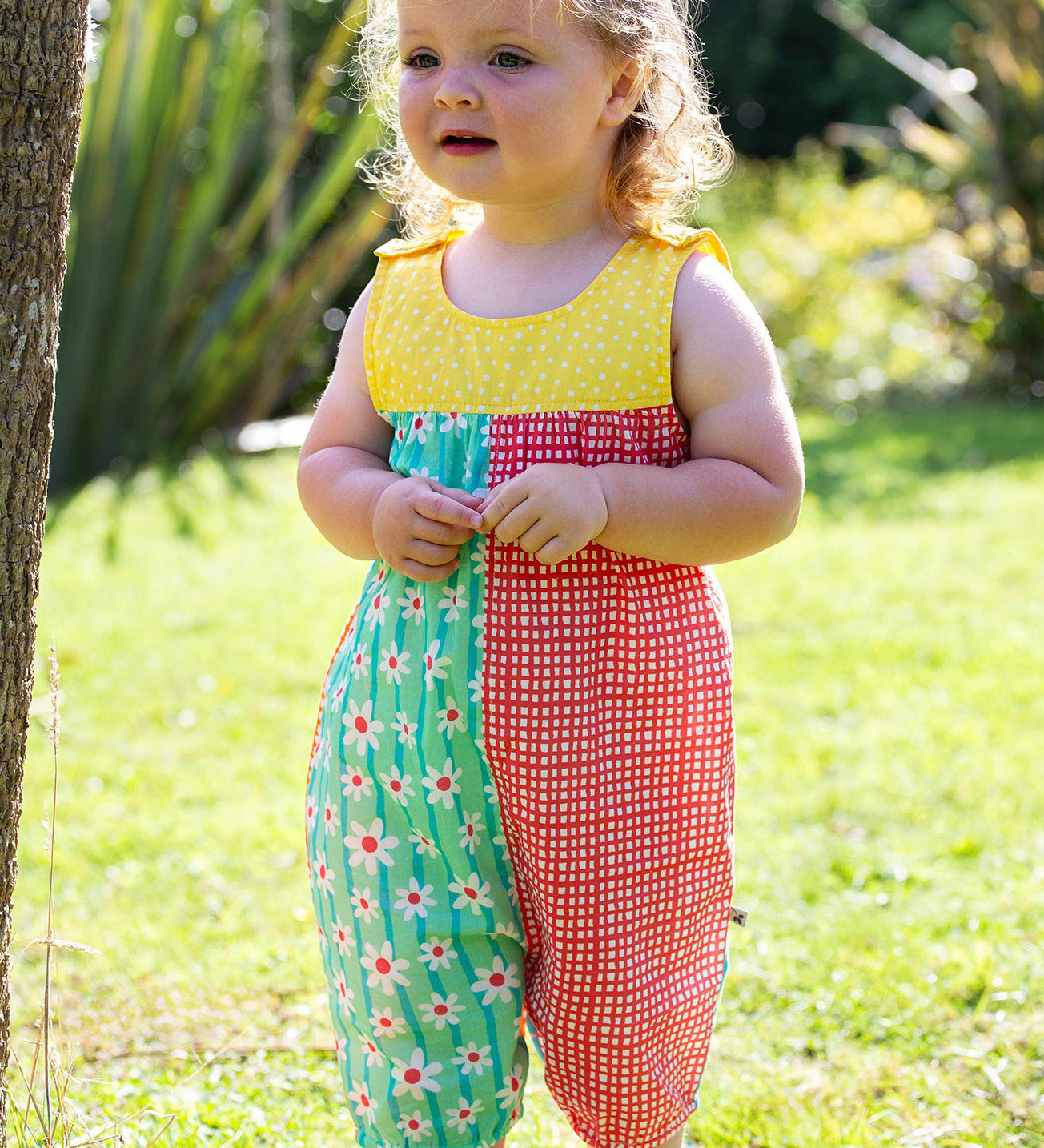 A toddler wearing Frugi Summer Printed Dungaree in the Rainbow Hotchpotch print, featuring the patchwork design and popper fastenings, standing on grass surrounded by plants.