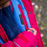A close up of a child pulling down the pink zip from the hidden side pocket on the inside of the Frugi 3 in 1 snapdragon/ navy  waterproof rambler coat showing the 'treasures & treats' patch stitched on the lilac post consumer recycled polyester fabric