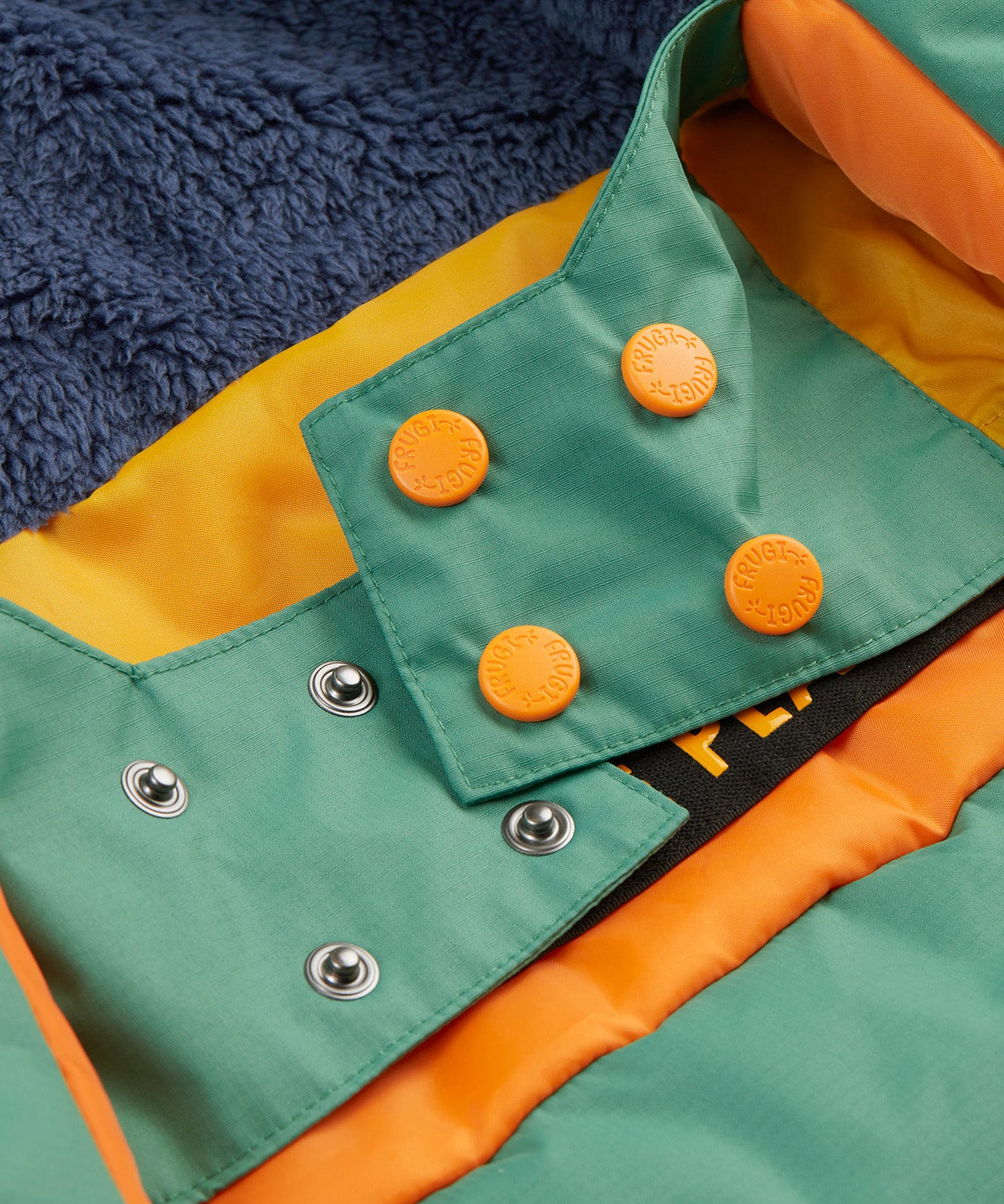 A close up of the Frugi kids snow and ski coat in green with a mountain scene print. Showing the thick navy fleece on the back and popper fasteners to adjust the size for a snug fit