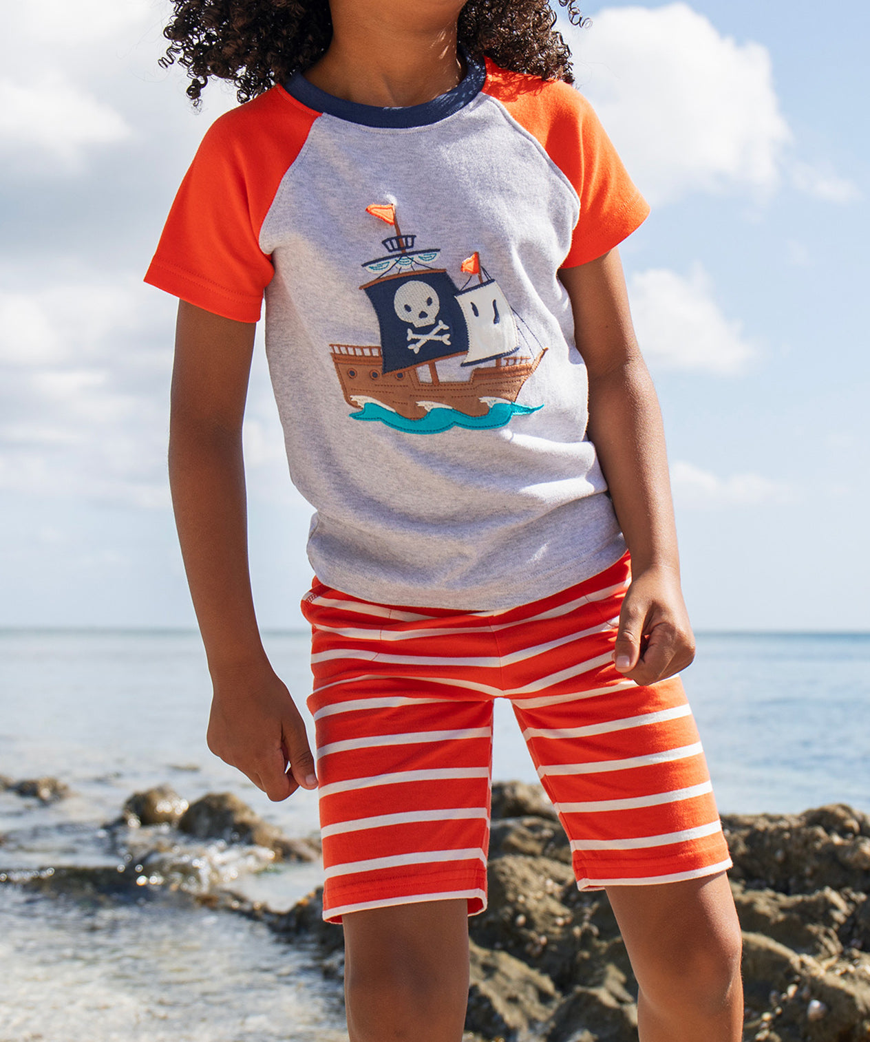 A young child wearing Frugi Stithian Shorts in Tiger Orange Breton, standing on a rock by the sea.