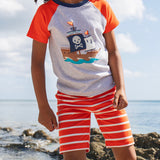 A young child wearing Frugi Stithian Shorts in Tiger Orange Breton, standing on a rock by the sea.