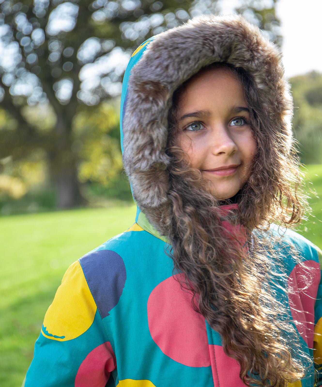 A child wearing the Frugi kids penhale super spot parka outside showing the furry hood detail and the big circles design on light blue organic cotton fabric.