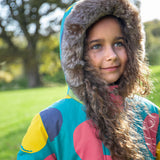 A child wearing the Frugi kids penhale super spot parka outside showing the furry hood detail and the big circles design on light blue organic cotton fabric.
