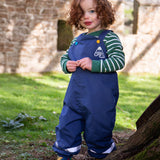 A toddler standing outside wearing the Frugi navy waterproof dungarees with reflective straps on the legs. Features a blue and yellow button fastener for the shoulder strap for a secure fit