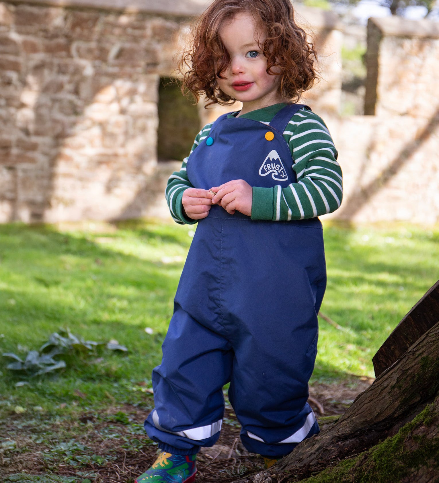 A toddler standing outside wearing the Frugi navy waterproof dungarees with reflective straps on the legs. Features a blue and yellow button fastener for the shoulder strap for a secure fit