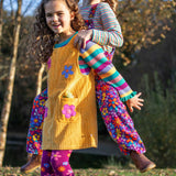 A child wearing the Frugi organic cotton Ginny yellow gold Corduroy Pinafore Dress with Flowers with retro  flower tights and a rainbow striped top underneath. This dress is a part of a wide range of organic cotton clothing for children available here at Babipur.