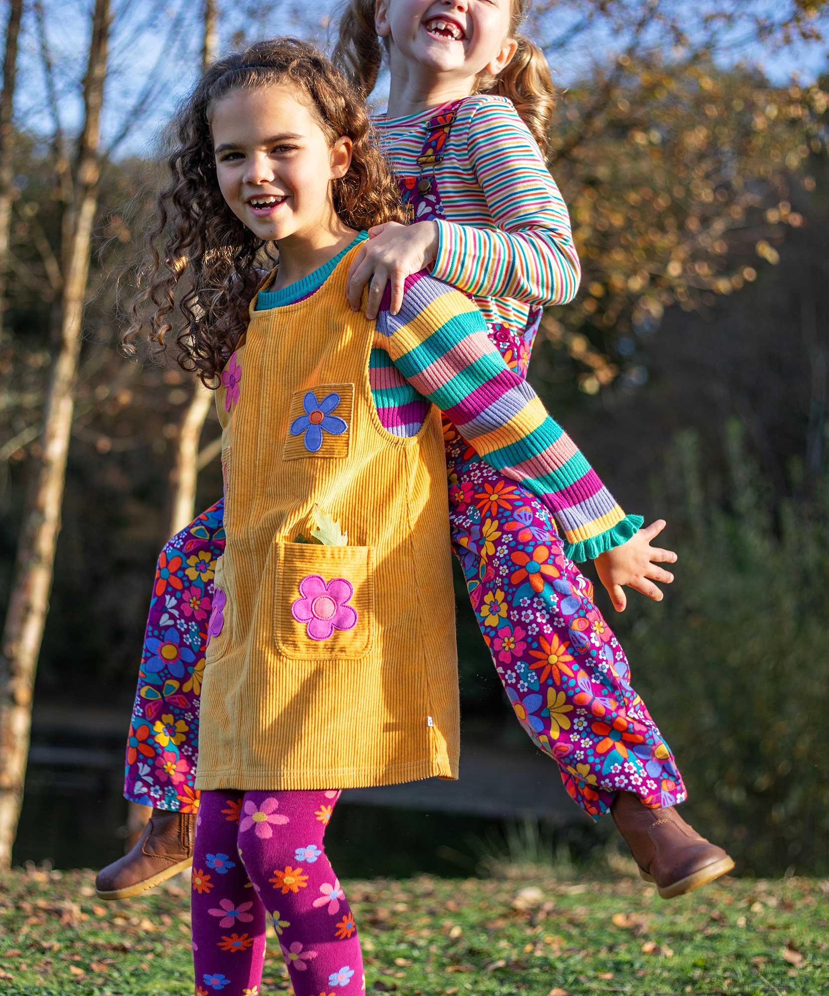 A child wearing the Frugi organic cotton Ginny yellow gold Corduroy Pinafore Dress with Flowers with retro  flower tights and a rainbow striped top underneath. This dress is a part of a wide range of organic cotton clothing for children available here at Babipur.