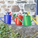 A group of 4 Glückskäfer 1L Metal Watering Cans all lined up on a stone wall outdoors. They are blue, red, green and yellow.
