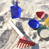 A Glückskäfer Metal Red Rake placed on the sand next to metal sand moulds. A blue round shovel can be seen in the background.