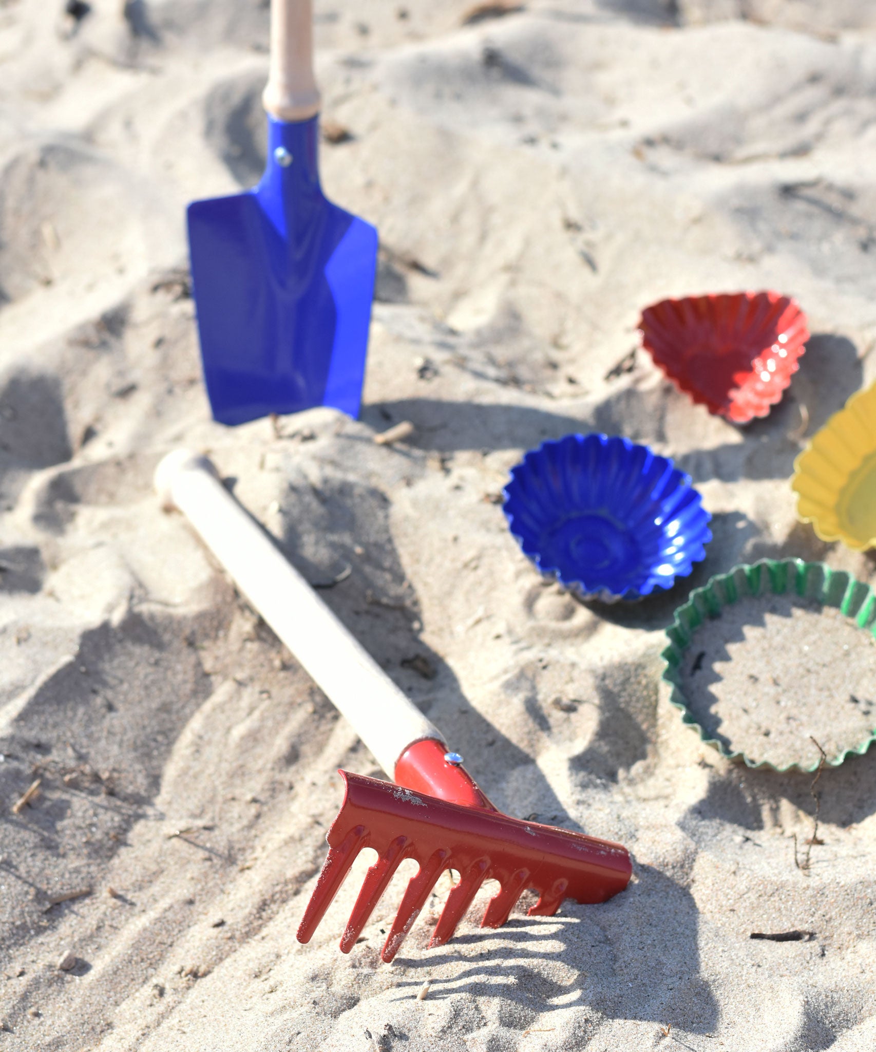 A Glückskäfer Metal Red Rake placed on the sand next to metal sand moulds. A blue round shovel can be seen in the background.