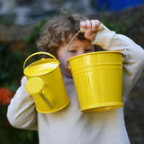 A child holding up a Glückskäfer yellow 1L Metal Watering can and a bucket.