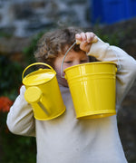 A child holding up a Glückskäfer yellow 1L Metal Watering can and a bucket.