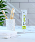 Green People Fennel & Propolis Natural Toothpaste on a marble surface in a bathroom next to a ceramic pot with bamboo toothbrushes.