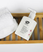 A bottle of Green People Manuka & Lemon Tea Tree Hand Wash placed on a bamboo bath shelf next to a fluffy white towel.