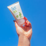 An adult's hand holding up a tube of Green People Children's Aloe Vera Aftersun Lotion against a clear blue sky.