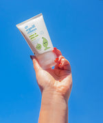 An adult's hand holding up a tube of Green People Children's Aloe Vera Aftersun Lotion against a clear blue sky.