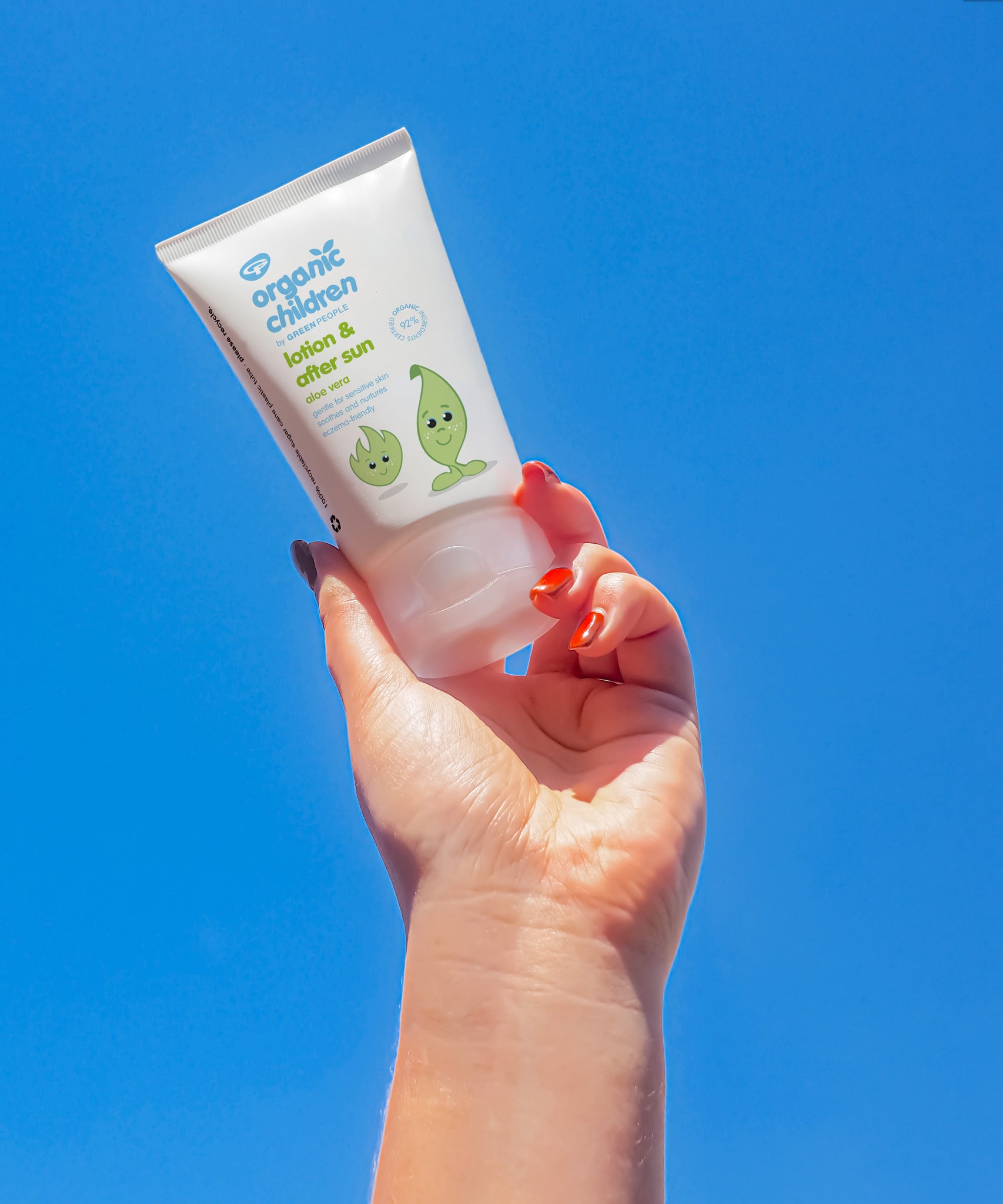 An adult's hand holding up a tube of Green People Children's Aloe Vera Aftersun Lotion against a clear blue sky.