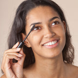 A person with dark hair applying the Green People Volumising Black Mascara to their eyelashes. The person is looking up and smiling.