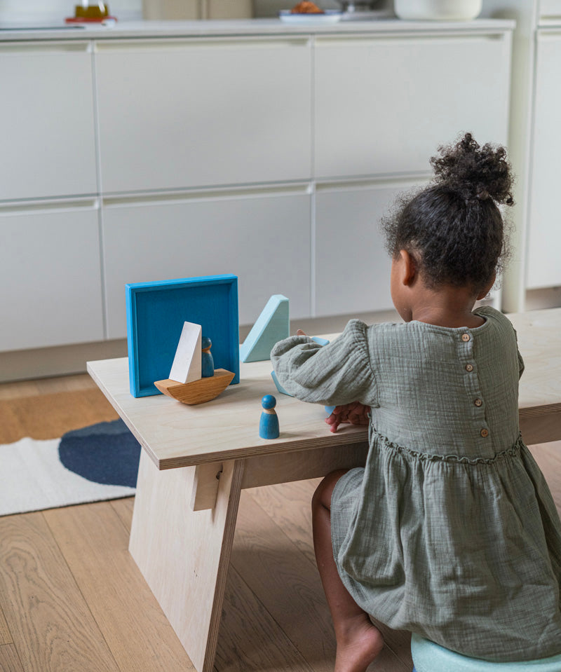 A child playing with the Grimm's blue boat Wooden Stacking Puzzle. The child has the blocks placed in a boat shape and has placing Grimm's friends wooden peg dolls on the boat. These Grimm's toys are part of a wide range of wooden toys available here at Babipur.