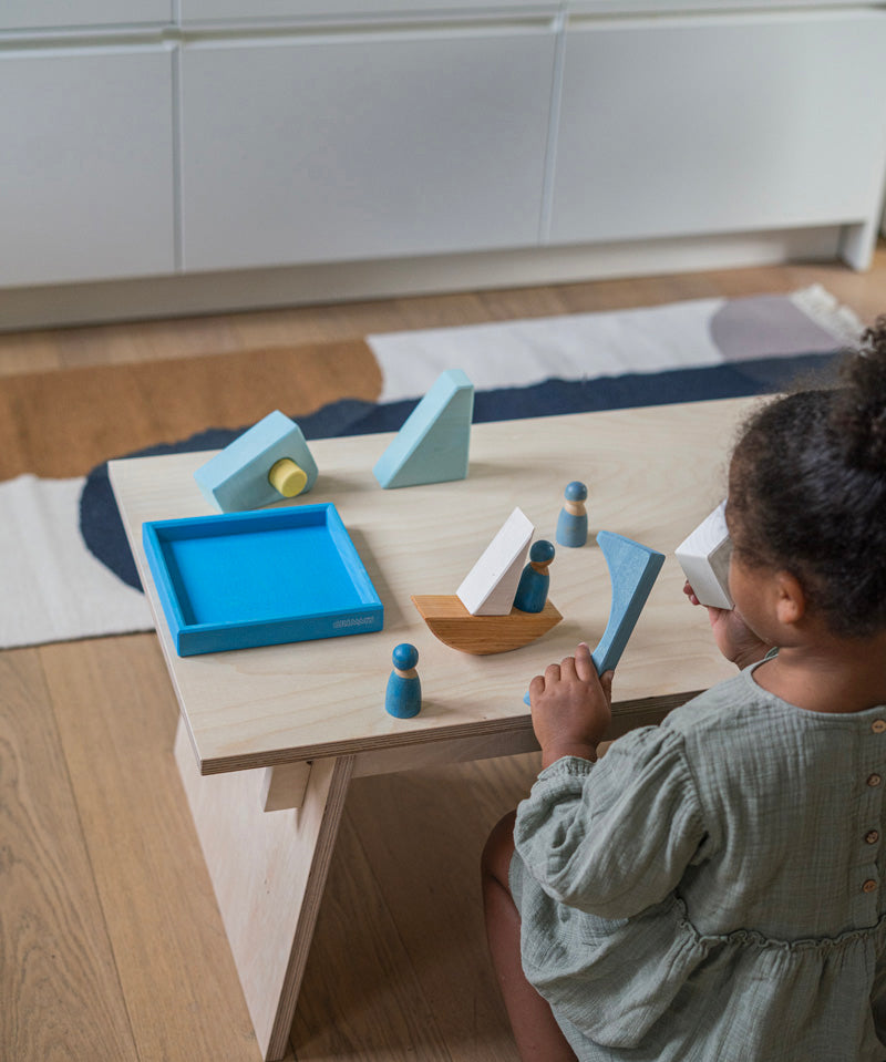 A child playing with the Grimm's blue boat Wooden Stacking Puzzle. The child has the blocks placed in a boat shape and has placing Grimm's friends wooden peg dolls on the boat. These Grimm's toys are part of a wide range of wooden toys available here at Babipur.