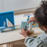 A child playing with the Grimm's blue boat Wooden Stacking Puzzle. The child has the blocks placed in a boat shape and is placing Grimm's friends wooden peg dolls on the boat. These Grimm's toys are part of a wide range of wooden toys available here at Babipur.
