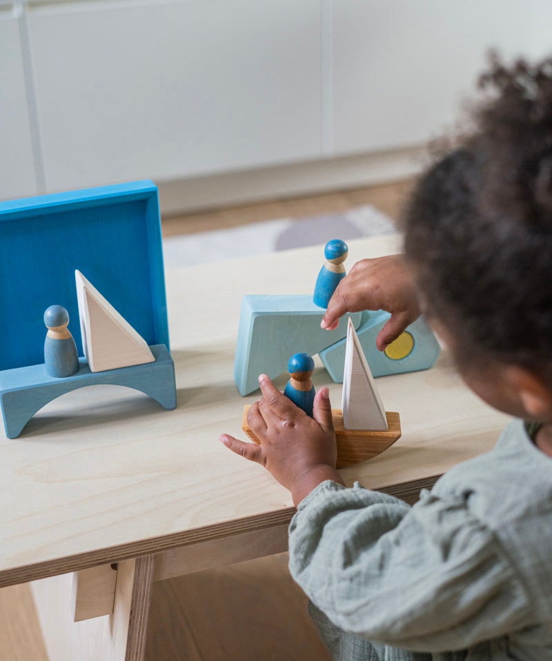 A child playing with the Grimm's blue boat Wooden Stacking Puzzle. The child has the blocks placed in a boat shape and is placing Grimm's friends wooden peg dolls on the boat. These Grimm's toys are part of a wide range of wooden toys available here at Babipur.