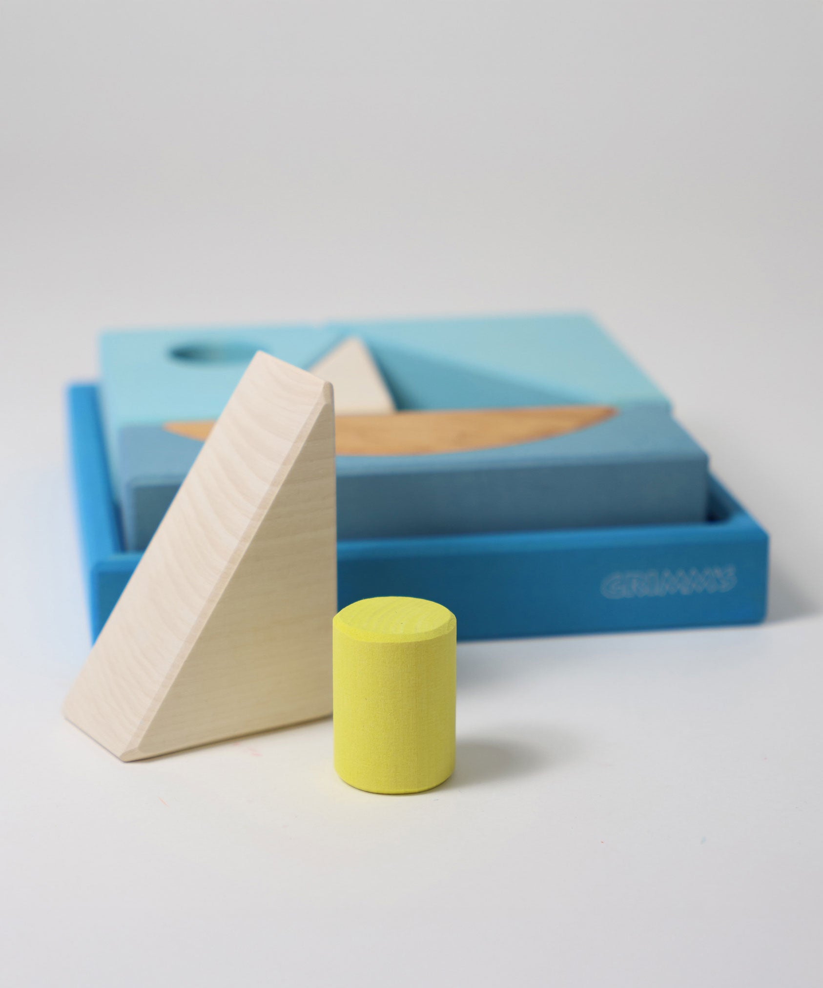 The white triangle and green cylinder wooden block from the Grimm's blue building set placed in front of the blue tray. These Grimm's wooden block sets are part of a wide range of wooden toys available here at Babipur.