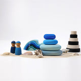 A set of 4 Grimm's wooden pebbles in a blue river colourway shown amongst a seaside themed small world play set up. There are two Grimm's friends besides teh stacked up blue pebbles surrounded by sand and shells. other wooden toys from Grimm's including waves and a monochrome stacker can be seen in the background.