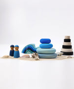 A set of 4 Grimm's wooden pebbles in a blue river colourway shown amongst a seaside themed small world play set up. There are two Grimm's friends besides teh stacked up blue pebbles surrounded by sand and shells. other wooden toys from Grimm's including waves and a monochrome stacker can be seen in the background.