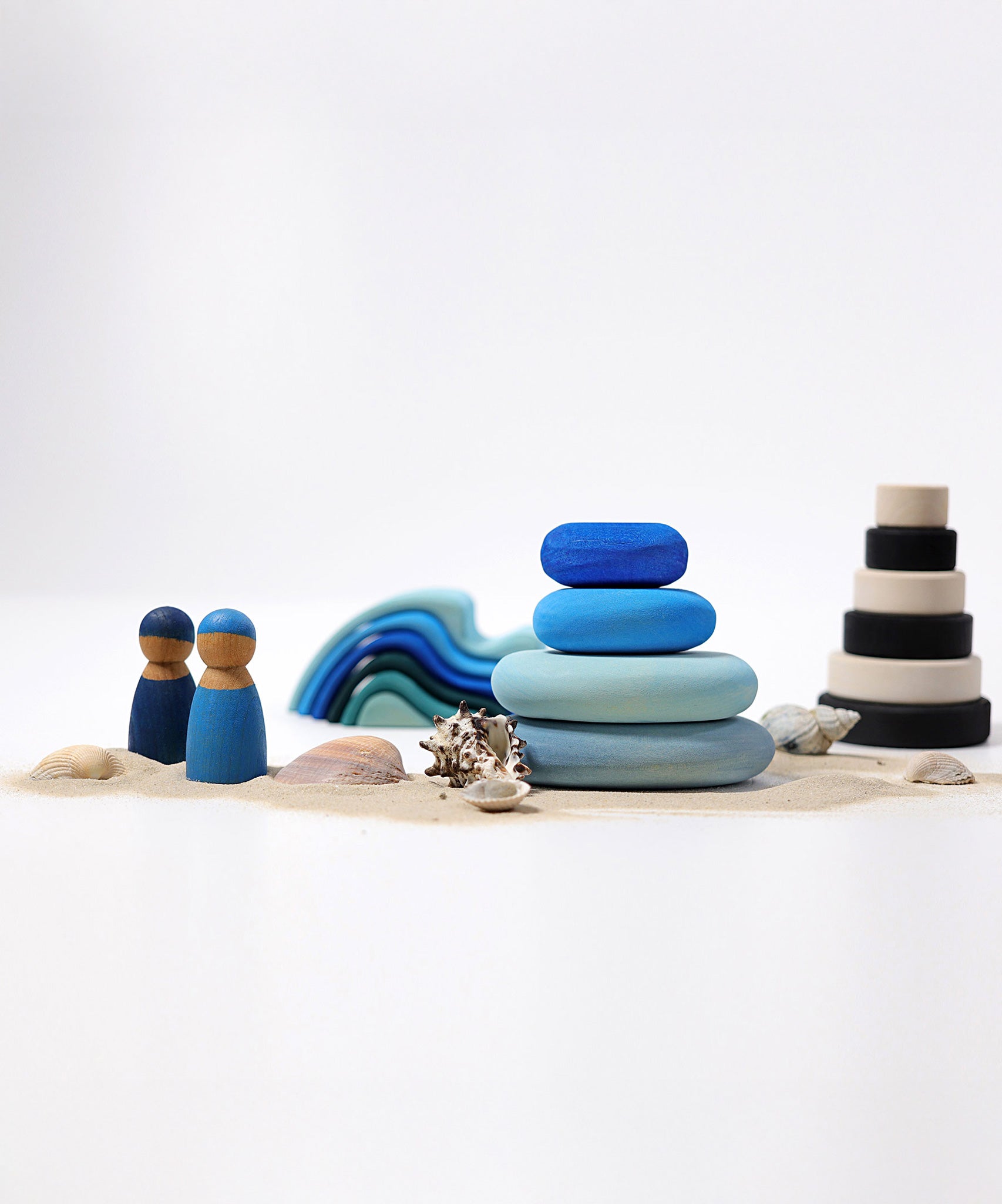 A set of 4 Grimm's wooden pebbles in a blue river colourway shown amongst a seaside themed small world play set up. There are two Grimm's friends besides teh stacked up blue pebbles surrounded by sand and shells. other wooden toys from Grimm's including waves and a monochrome stacker can be seen in the background.
