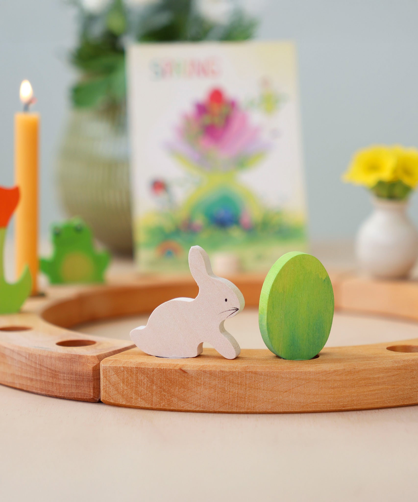Grimm's Easter Decorative Figure Set featuring a white bunny rabbit a Batik Green coloured eggs. The figures are shown on a natural wooden celebration ring. Other decorative figures can be seen placed on the ring in the background.