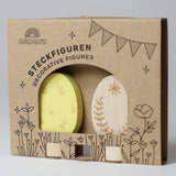 Grimm's Ornamental Duo Easter Eggs Decorative Figure Set. One egg is left natural withe floral themed outline details, the other painted in a light yellow with leaf outline details. The eggs are shown in their cardboard packaging sleeve.