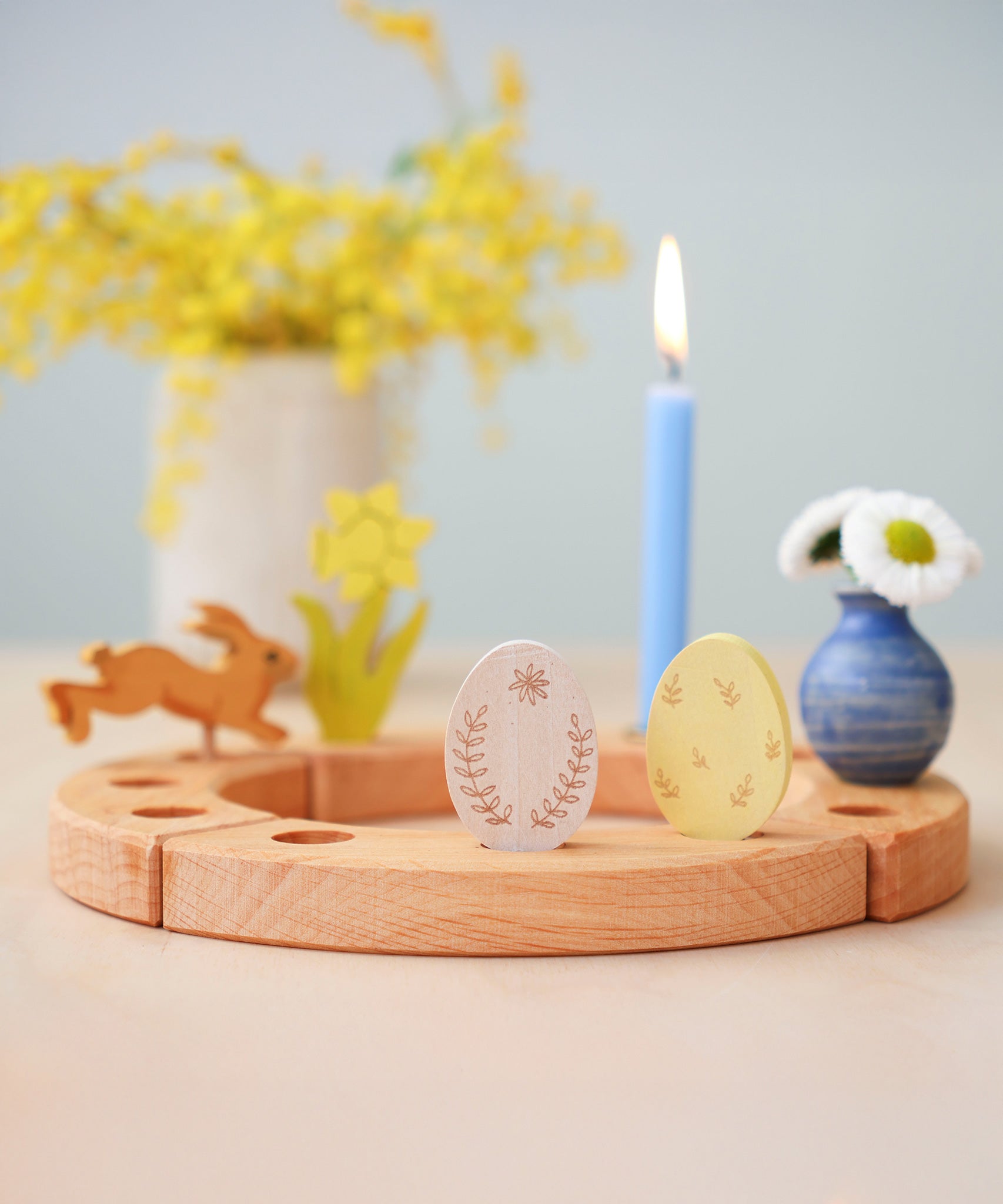 Grimm's Ornamental Duo Easter Eggs Decorative Figure Set. The eggs are placed on a natural wooden Grimm's celebration ring. Other decorative figures and a blue coloured candle can be seen in the background.