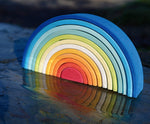 The Grimm's Gwawr Sunrise 12 Piece Rainbow shown on a wet slate surface outdoors. The reflection of the rainbow can be seen in the water,.