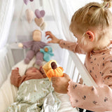 An older child is looking over the side of a white cot with a baby inside. The older child is holding the Grimm's Lavender Doll Sun in their hand. The image also shows other Grimm's Doll at the head of the cot with a pink and purple fabric heart mobile above the cot