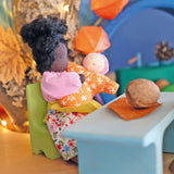 Grimm's Mrs Ebony is holding Baby Mia, whilst sat on a green wooden chair. The background has an autumnal theme with brown and orange colours, leaves and pinecones