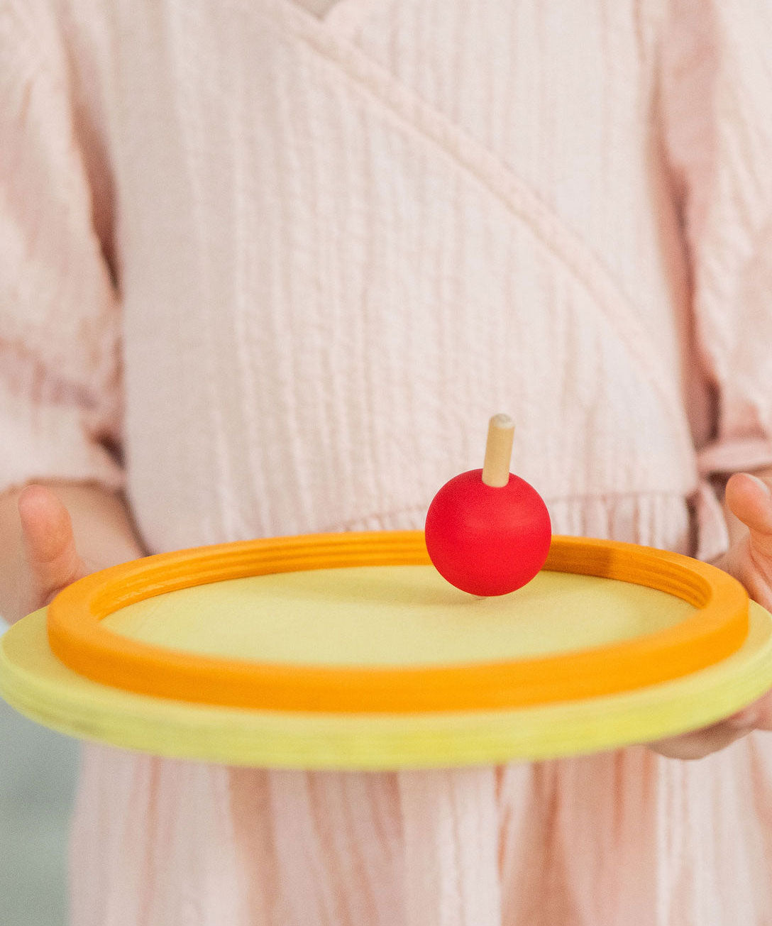 A child holding a red coloured rainbow spinning top on a circular wooden base. These Grimm's toys are part of a wide range of wooden toys available here at Babipur.