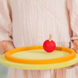 A child holding a red coloured rainbow spinning top on a circular wooden base. These Grimm's toys are part of a wide range of wooden toys available here at Babipur.