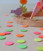 A close of the Grimm's Neon Confetti Dots scattered next to a celebration ring with a singing birds and neon pink cone sitting on top of the ring