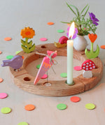 A Grimms wooden neon confetti dots scattered next to a celebration ring with different decorative figures on top including a purple singing bird, a orange flower, pink neon birthday cone, mushroom, snail, white vase and a beeswax candle.