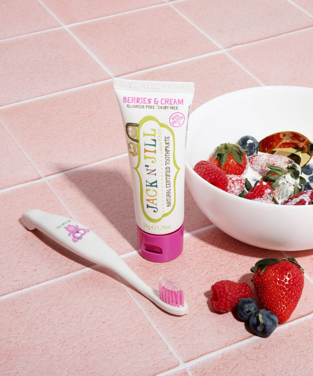 A 50g tube of Jack N' Jill berries and cream flavoured fluoride free kid's toothpaste placed on pink tiles next to a Jack N' Jill toothbrush. A bowl full of berries and cream can be seen to the side.