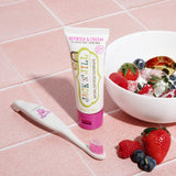 A 50g tube of Jack N' Jill berries and cream flavoured fluoride free kid's toothpaste placed on pink tiles next to a Jack N' Jill toothbrush. A bowl full of berries and cream can be seen to the side.
